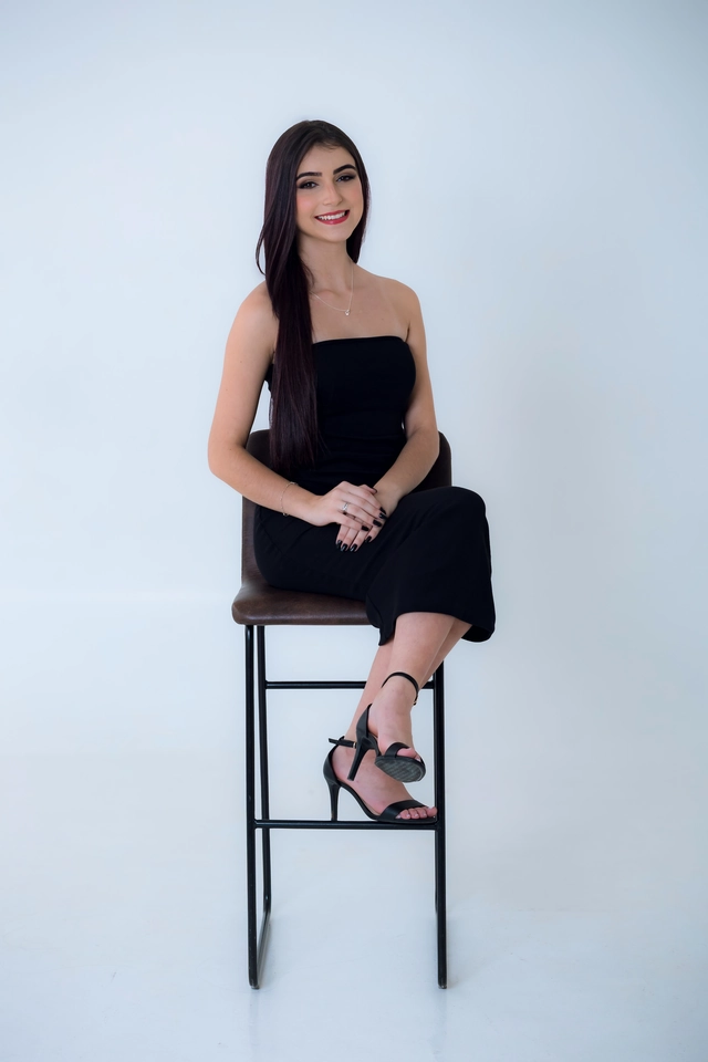 Formanda sentada no banco, vestido preto, mãos cruzadas no colo