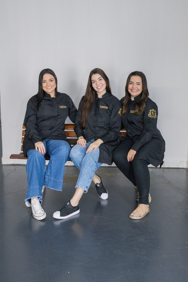 Três formandas de Odontologia sentadas num banco, com jaleco preto bordado