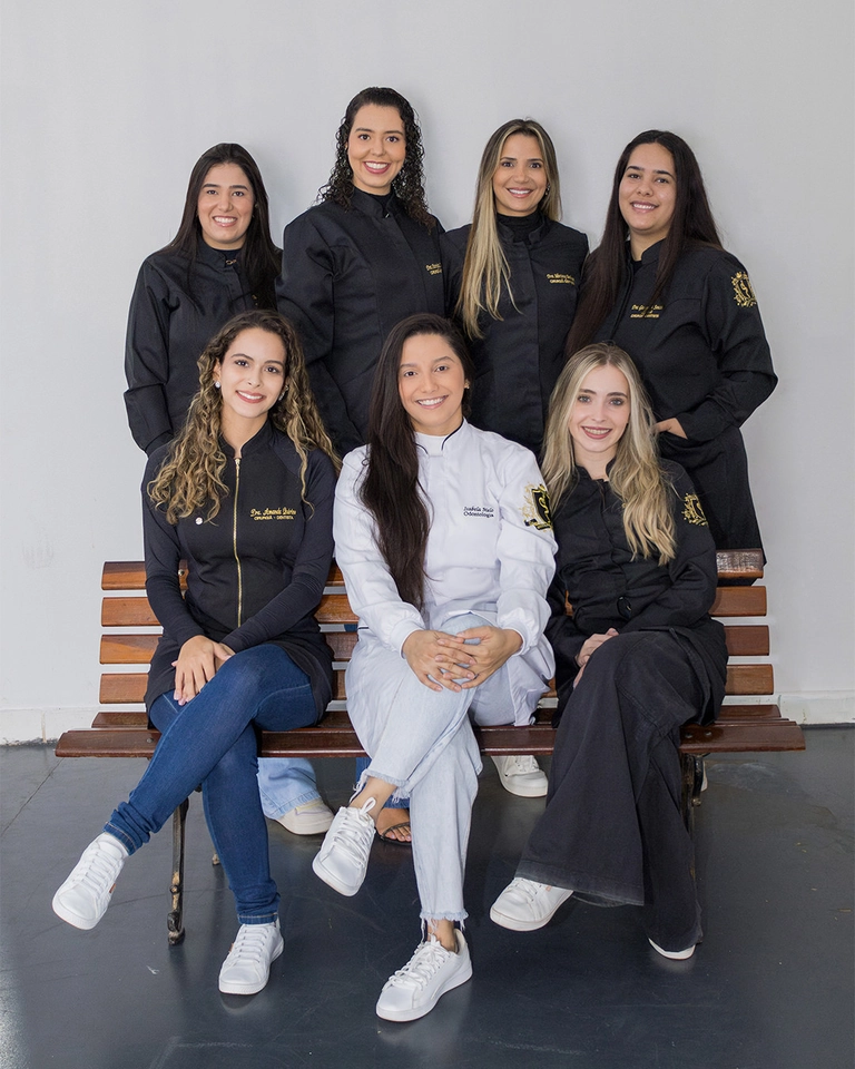 Sete formandos de Odontologia reunidos para retrato de grupo, com uma formanda de jaleco branco ao centro