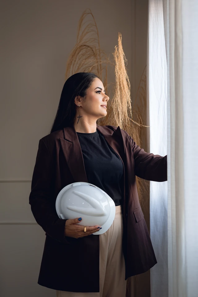Formanda observando pela janela, segurando o capacete de engenharia